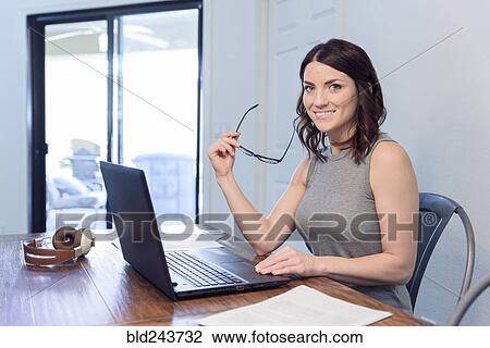 Stock Image - Smiling Caucasian woman holding eyeglasses using laptop. Fotosearch