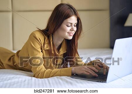 Stock Image - Smiling Caucasian woman laying on bed using laptop. Fotosearch
