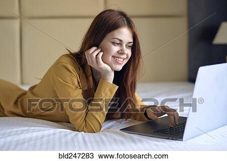 Stock Image - Smiling Caucasian woman laying on bed using laptop. Fotosearch