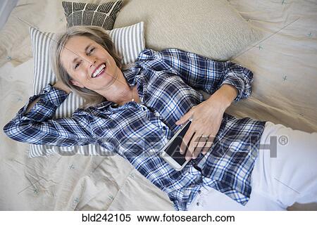 Stock Photography - Smiling Caucasian woman laying on bed holding cell phone. Fotosearch