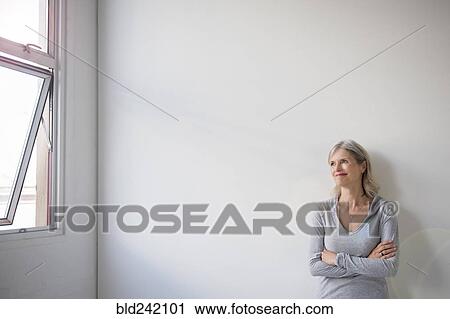 Stock Image - Smiling Caucasian woman leaning on wall near window. Fotosearch