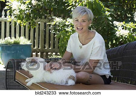 Stock Image - Smiling Caucasian woman petting dog on sofa outdoors. Fotosearch