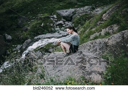 Stock Image - Smiling Caucasian woman sitting on rock on hill. Fotosearch