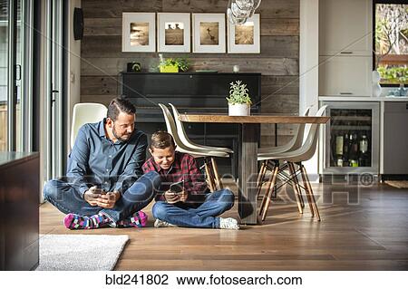 Stock Image - Smiling father and son sitting on floor texting on cell phones. Fotosearch