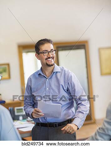 Picture - Smiling Hispanic businessman holding paperwork in office. Fotosearch