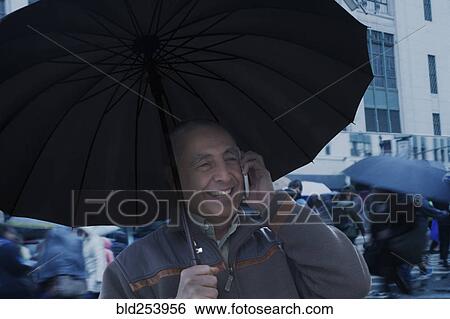 Stock Photograph - Smiling Hispanic man holding umbrella and talking on cell phone. Fotosearch