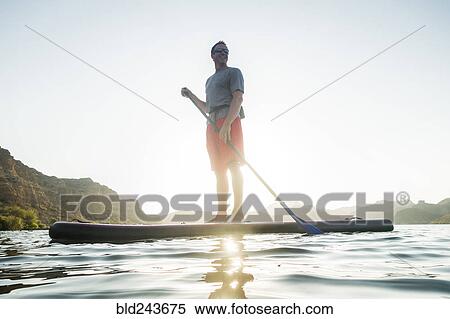 Stock Photography - Smiling Hispanic man standing on paddleboard in river. Fotosearch