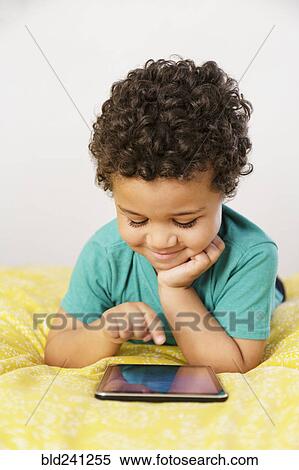Smiling Mixed Race boy laying on bed using digital tablet View Large Photo Image Stock Photography - Smiling Mixed Race boy laying on bed using digital tablet. Fotosearch