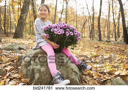 Stock Image - Smiling Mixed Race girls sitting on rock in autumn holding flowers. Fotosearch