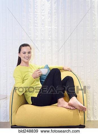 Stock Photo - Smiling mixed race woman drinking coffee in yellow chair. Fotosearch