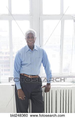 Stock Image - Smiling older Black man leaning on radiator near window. Fotosearch