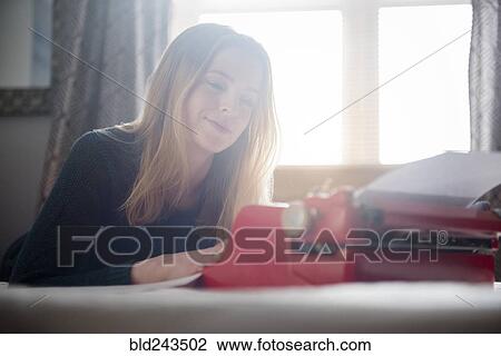 Stock Image - Smiling woman laying on bed typing on typewriter. Fotosearch