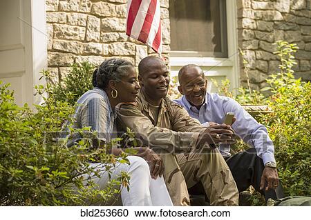 Stock Image - Soldier sitting on front stoop with parents and texting on cell phone. Fotosearch