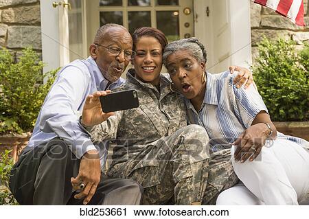 Stock Image - Soldier sitting on front stoop with parents and posing for cell phone selfie. Fotosearch