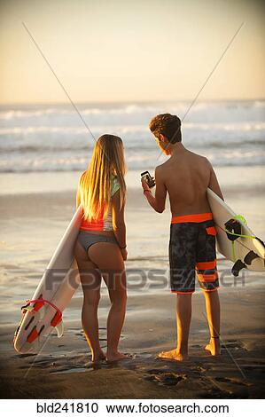 Stock Image - Teenage boy and girl holding surfboards texting on cell phone. Fotosearch