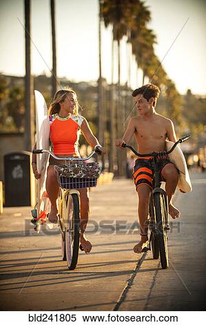 Stock Photography - Teenage boy and girl riding bicycles carrying surfboards. Fotosearch