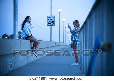 Stock Image - Teenage girl photographing friend sitting on wall. Fotosearch