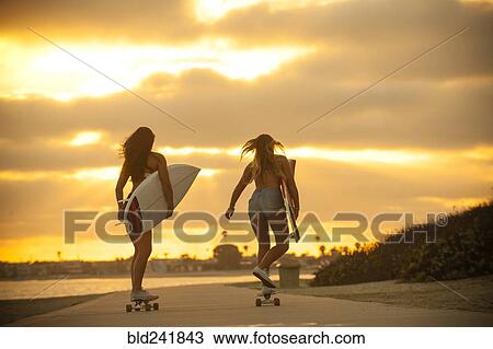 Stock Image - Teenage girls carrying surfboards on skateboards. Fotosearch