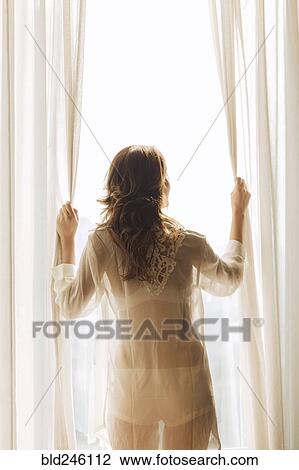 Stock Image - Thai woman wearing sheer clothing opening curtains at window. Fotosearch