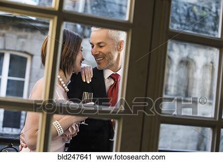 Stock Image - Well-dressed Caucasian couple drinking champagne behind window. Fotosearch
