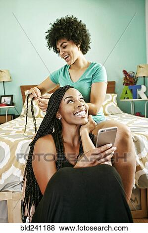 Woman braiding hair of friend holding cell phone in bedroom View Large Photo Image Stock Photo - Woman braiding hair of friend holding cell phone in bedroom. Fotosearch
