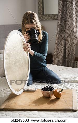Stock Image - Woman kneeling on bed holding reflector photographing fruit. Fotosearch