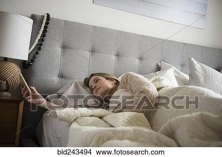 Picture - Woman laying in bed texting on cell phone. Fotosearch