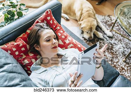 Stock Image - Woman laying on sofa near dog using digital tablet. Fotosearch