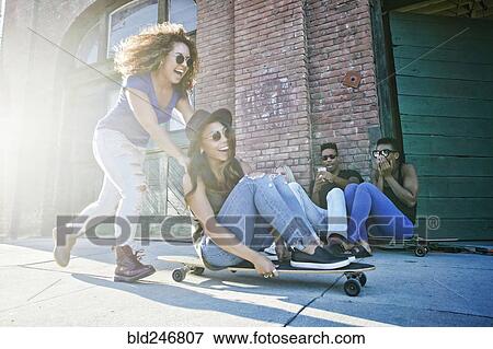 Stock Photo - Woman pushing friend sitting on skateboard in city. Fotosearch