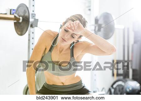 Stock Image - Woman resting in gymnasium wiping sweat from forehead. Fotosearch