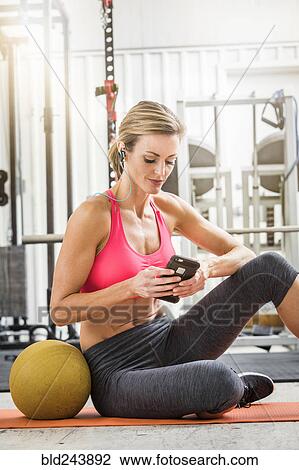 Stock Image - Woman sitting with heavy ball in gymnasium listening to cell phone. Fotosearch