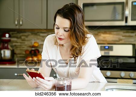 Stock Photograph - Woman texting on cell phone in domestic kitchen. Fotosearch