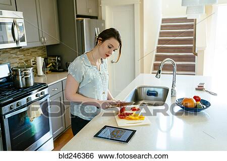 Stock Photograph - Woman using digital tablet and chopping food in domestic kitchen. Fotosearch