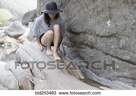 Stock Image - Woman wearing sun hat crouching on rock. Fotosearch