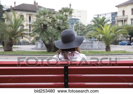 Stock Image - Woman wearing sun hat sitting on red bench. Fotosearch