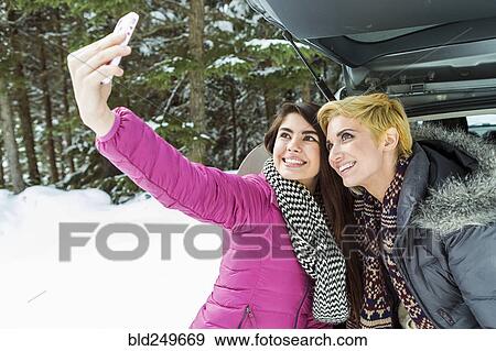 Stock Photo - Women at car posing for cell phone selfie in winter. Fotosearch
