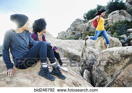 Stock Image - Women watching friend jumping on rock formation. Fotosearch