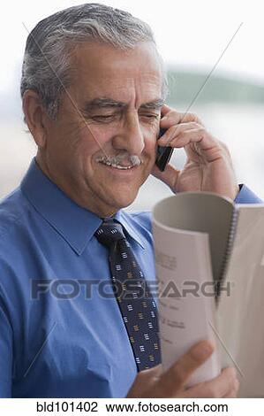 Stock Image - Hispanic businessman talking on cell phone and reading report. Fotosearch