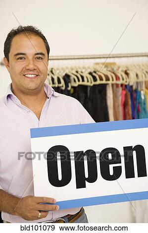 Stock Photo - Hispanic man holding open sign in clothing store. Fotosearch