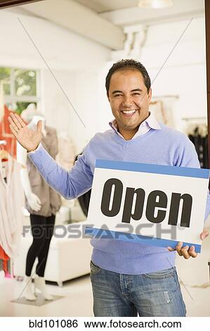 Stock Photograph - Hispanic man holding open sign in clothing store. Fotosearch