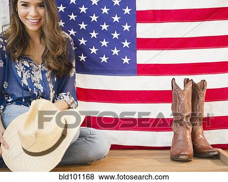 Stock Photo - Hispanic woman holding cowboy hat and sitting next to American flag. Fotosearch