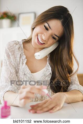Stock Image - Hispanic woman talking on phone and painting fingernails. Fotosearch