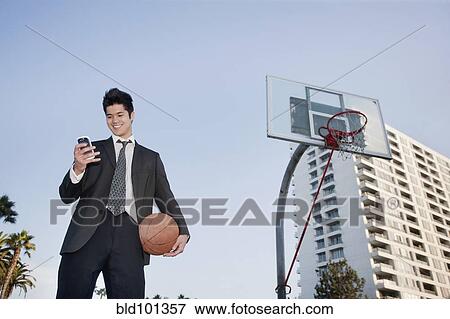 Stock Photo - Mixed race businessman holding basketball and text messaging on cell phone. Fotosearch