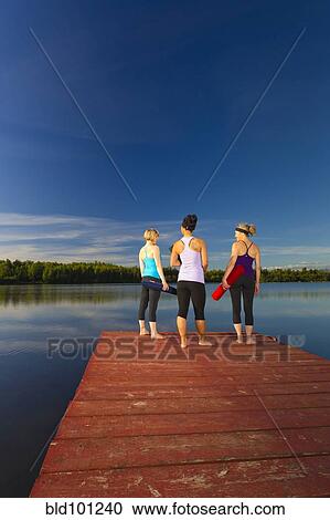 Stock Image - Women holding yoga mats on lake pier. Fotosearch