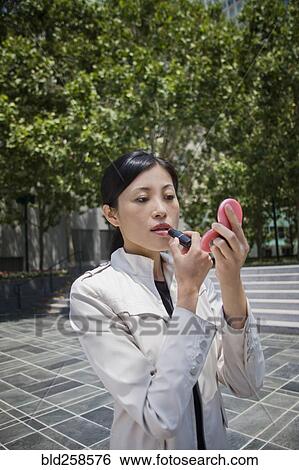 Stock Photograph - Asian woman applying lipstick outdoors. Fotosearch