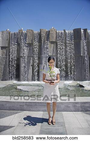 Stock Image - Asian woman holding seedling near fountain. Fotosearch