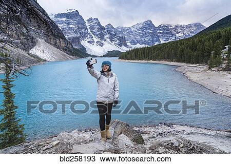 Stock Image - Asian woman posing for cell phone selfie at mountain lake. Fotosearch