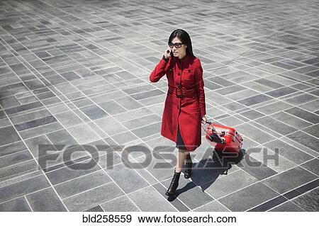 Stock Photo - Asian woman pulling rolling suitcase and talking on cell phone. Fotosearch