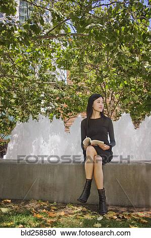 Stock Image - Asian woman sitting outdoors on concrete wall reading book. Fotosearch