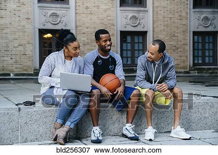 Stock Photo - Black friends sitting on concrete with basketball and laptop. Fotosearch